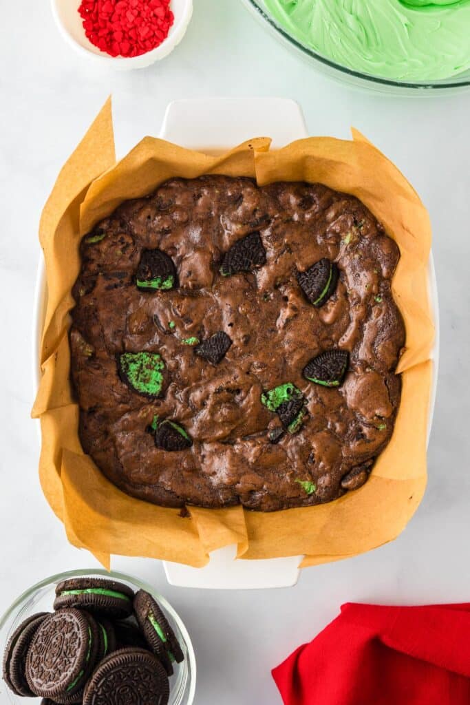 A pan of cooked chocolate Grinch brownies with chunks of mint Oreo cookies with more cookies, red heart sprinkles and green frosting in a bowl nearby on the counter.