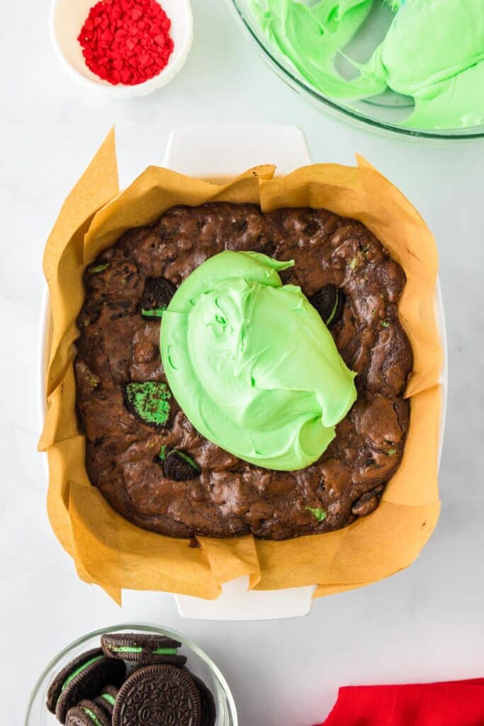 A pan of chocolate Grinch brownies with chunks of mint Oreo cookies being frosted with green frosting.