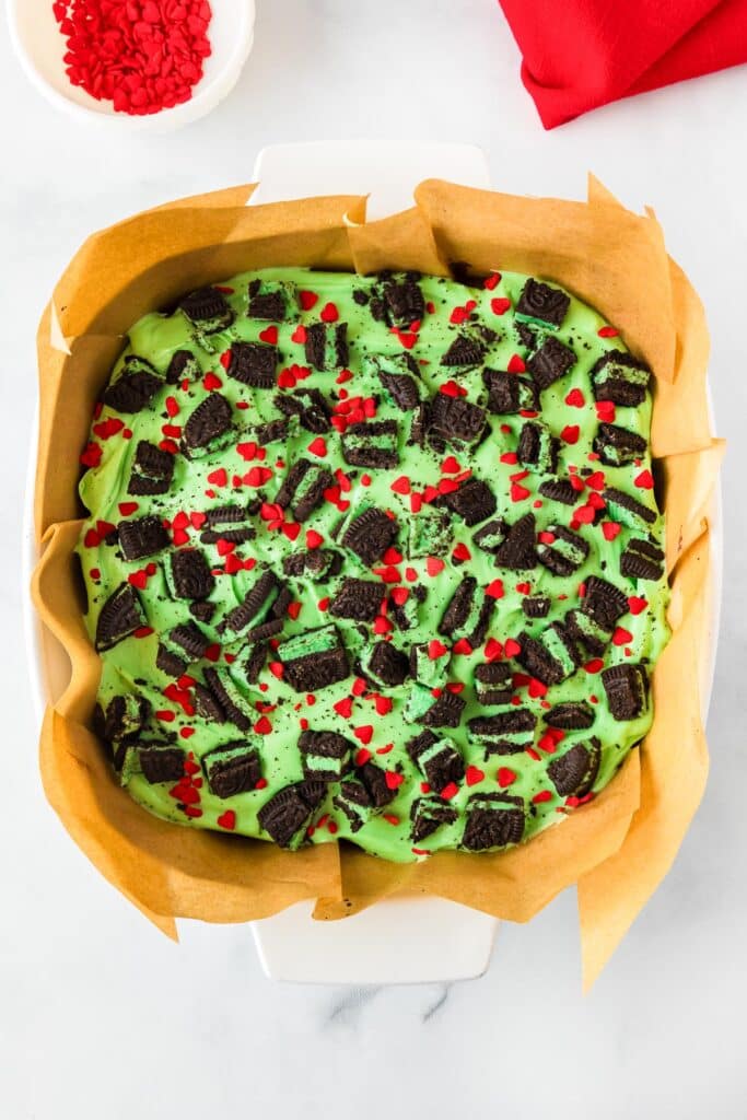 A pan of chocolate Grinch brownies topped with green frosting, chunks of mint Oreo cookies and red heart sprinkles.