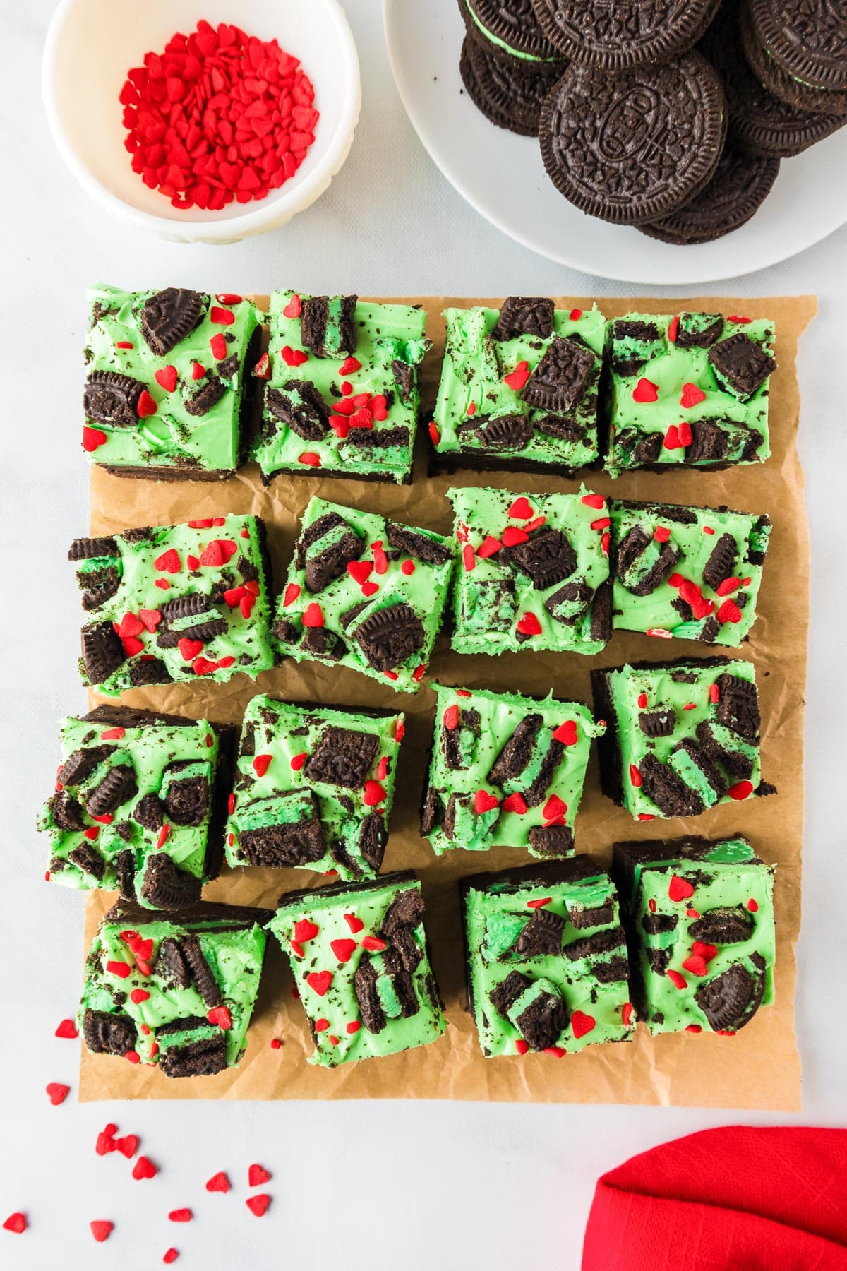 Green-frosted brownies GRinch brownies topped with crushed chocolate cookies and red heart sprinkles, arranged on parchment paper after slicing.