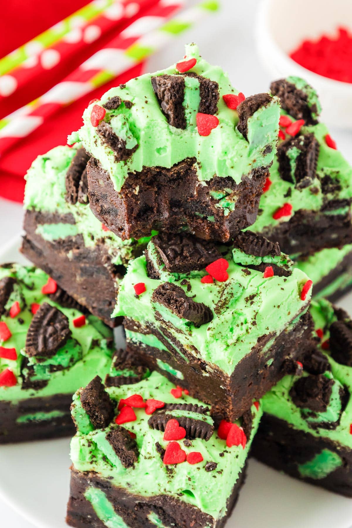 Stack of chocolate Grinch brownies topped with green frosting, crushed mint Oreo cookies, and red heart-shaped sprinkles on a plate with the top brownie missing a bite.