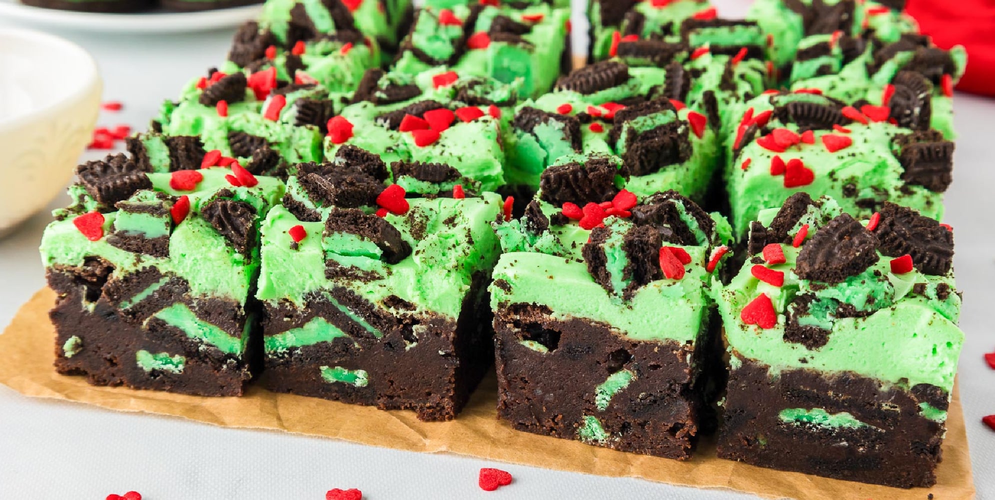 Mint chocolate Grinch brownies topped with green frosting, crushed mint Oreo cookies, and red heart sprinkles, cut into squares on a parchment paper.