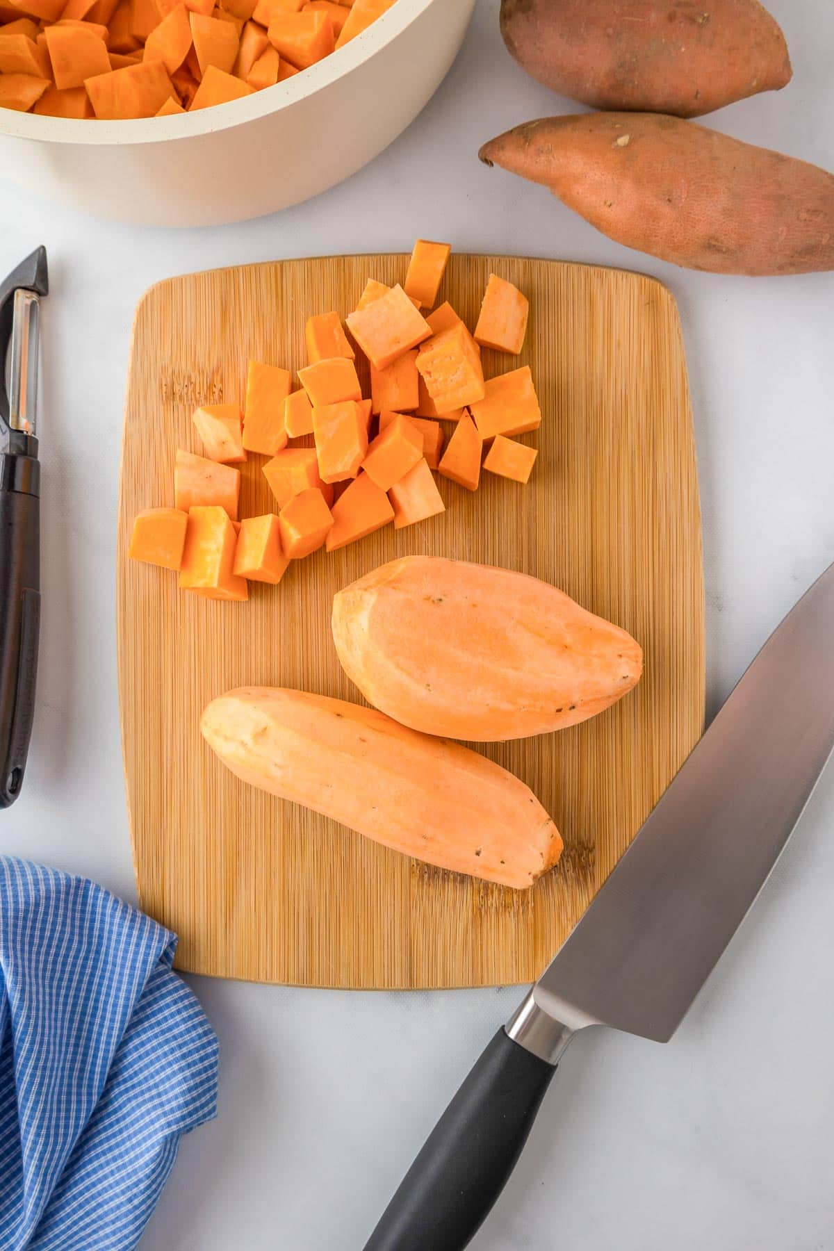 Cubed sweet potatoes for mashed sweet potatoes on a wooden cutting board with a knife, peeler, whole sweet potatoes, and a blue cloth nearby.