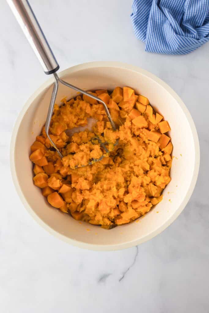 A bowl of sweet potatoes being mashed with a potato masher on a counter.