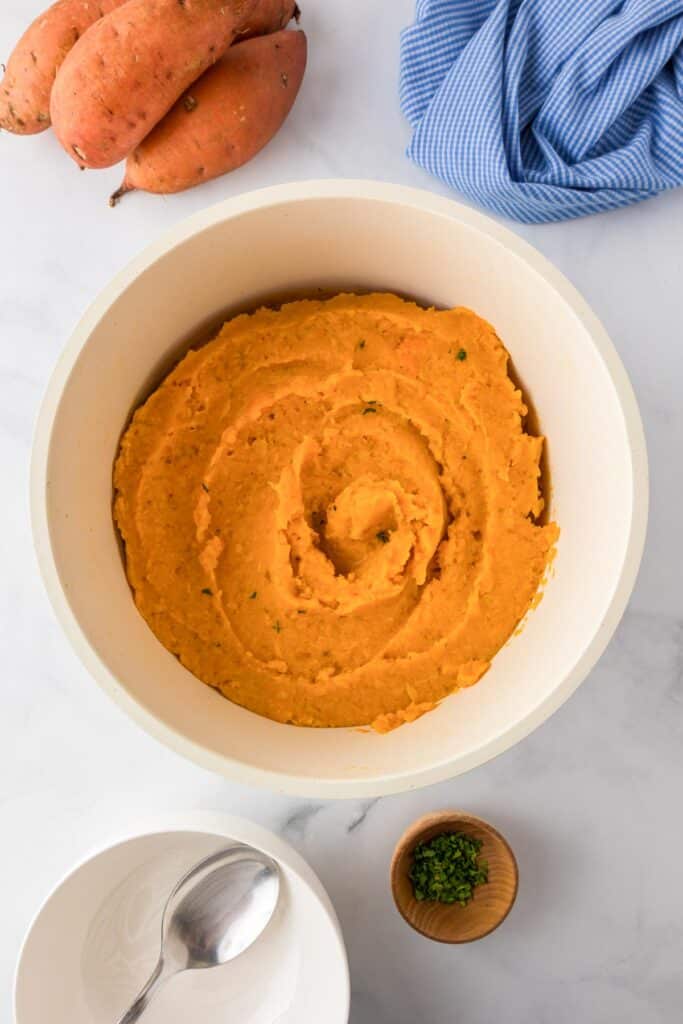 A bowl of mashed sweet potatoes in a large mixing bowl with a spoon, bowl, and parsley on the counter nearby.