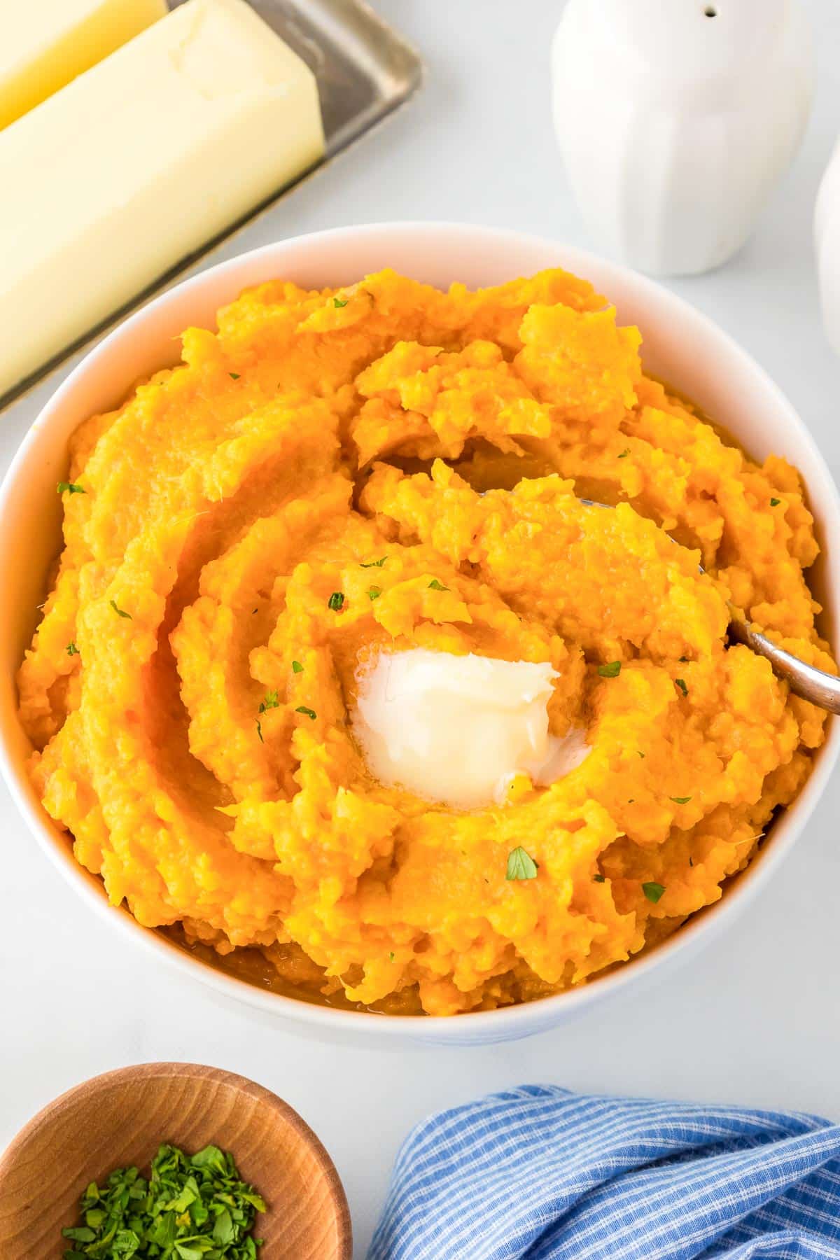 A bowl of mashed sweet potatoes with a pat of melted butter and as spoon scooping. More items nearby on the table such as salt, pepper, a stick of butter and more fresh herbs.