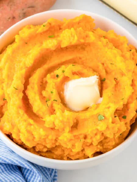 A bowl of maple mashed sweet potatoes in a bowl in a swirl pattern with a pat of melted butter on top on a table.