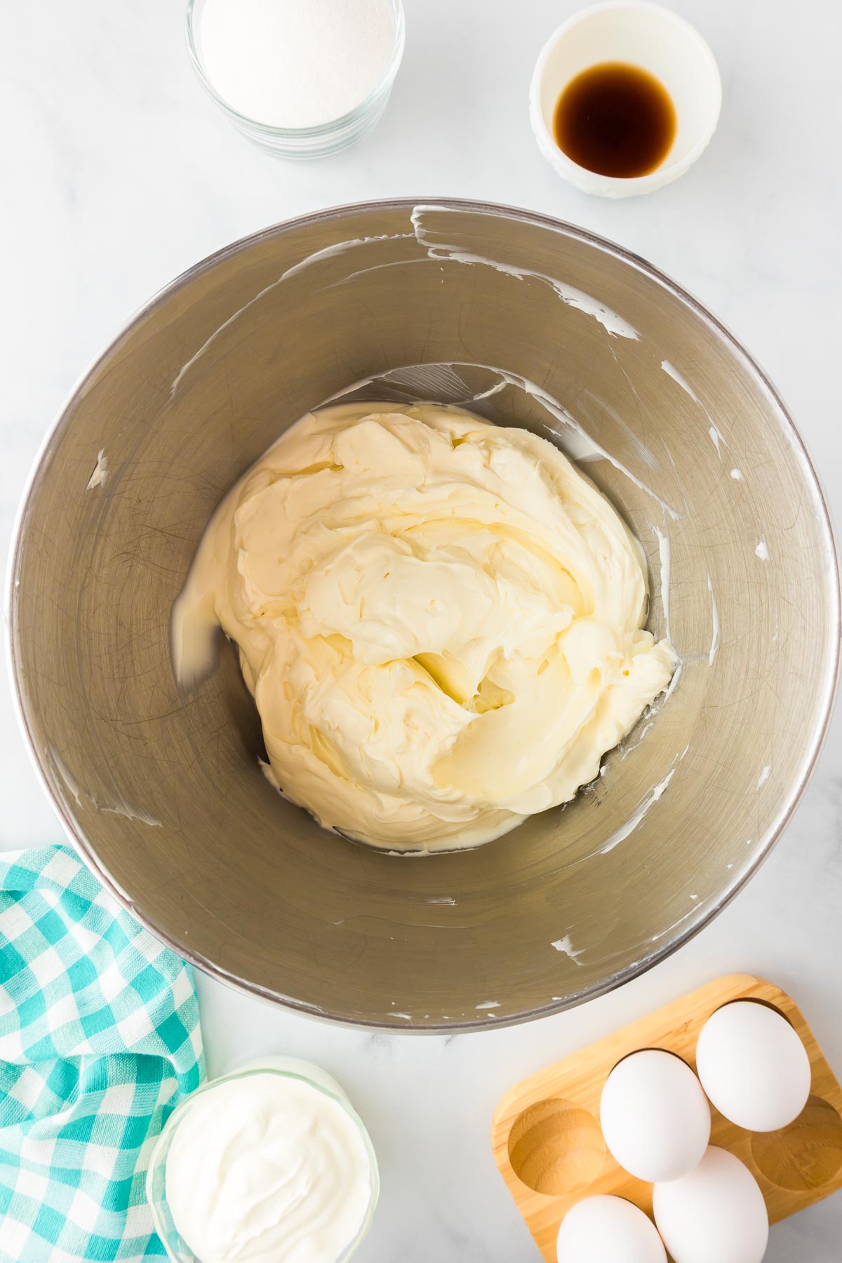 A mixing bowl with cream cheese that has been whipped with bowls of vanilla, sugar, eggs and sour cream nearby.