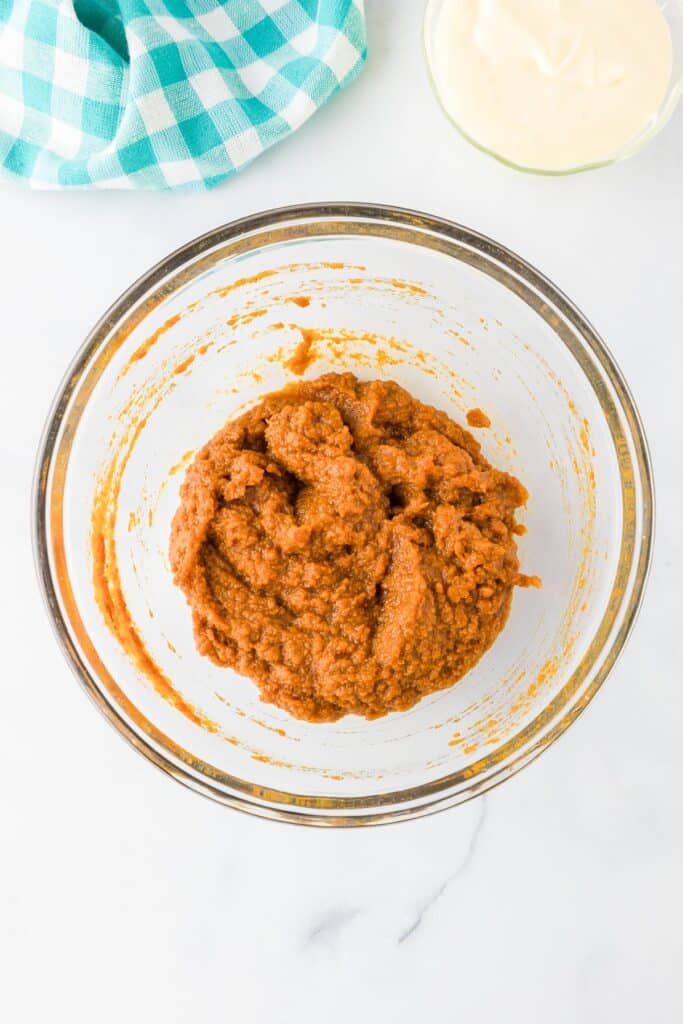 A glass bowl with a pumpkin puree mixture with a bowl of cheesecake filling nearby.