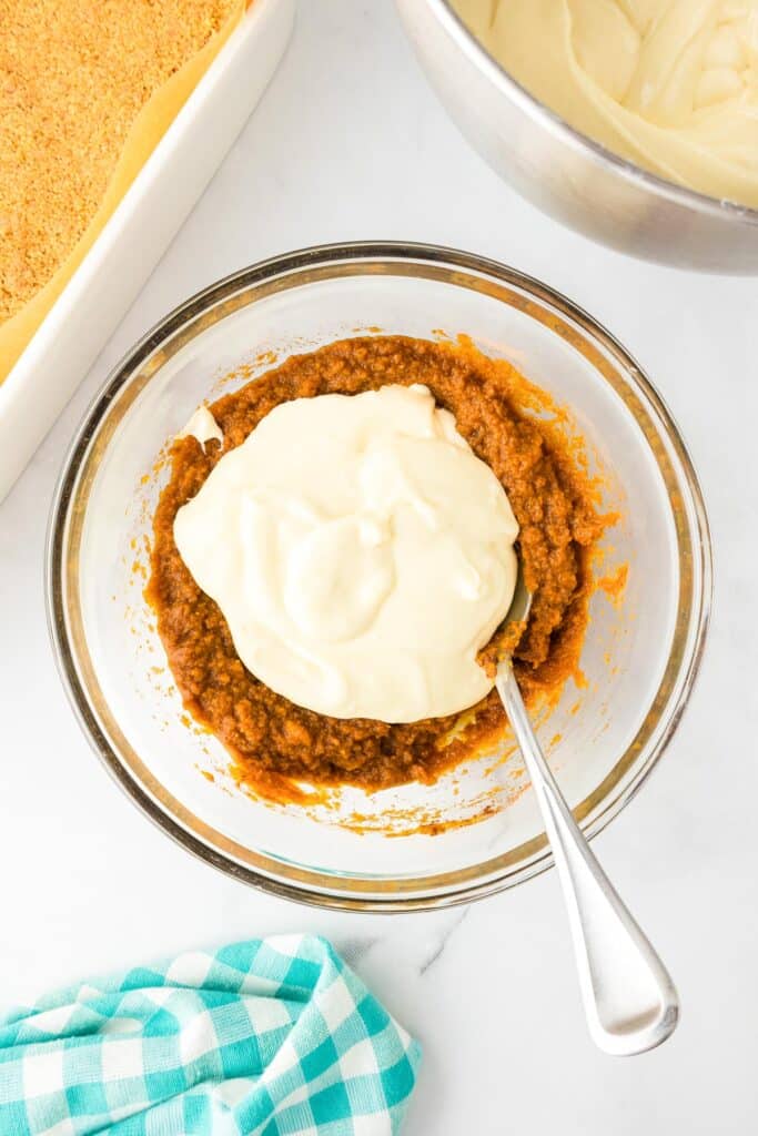 Pumpkin filling in a large bow being mixed with some of the cheesecake filling with more cheesecake filling in a bowl nearby and a pan full of graham cracker crust.