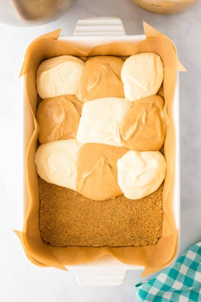 A baking dish lined with parchment paper filled with a graham cracker crust with large alternating dollops of cheesecake and pumpkin filling halfway down the pan.