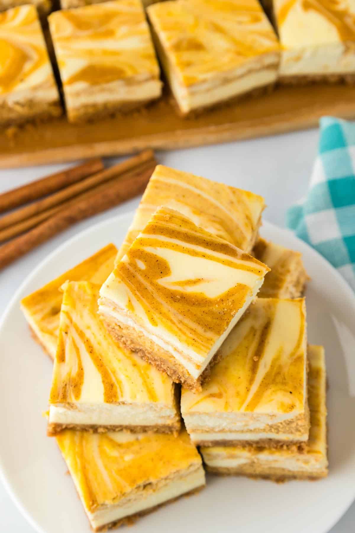 A plate stacked with pumpkin swirl cheesecake bars with more cheesecake bars lined up on a cutting board in the background.