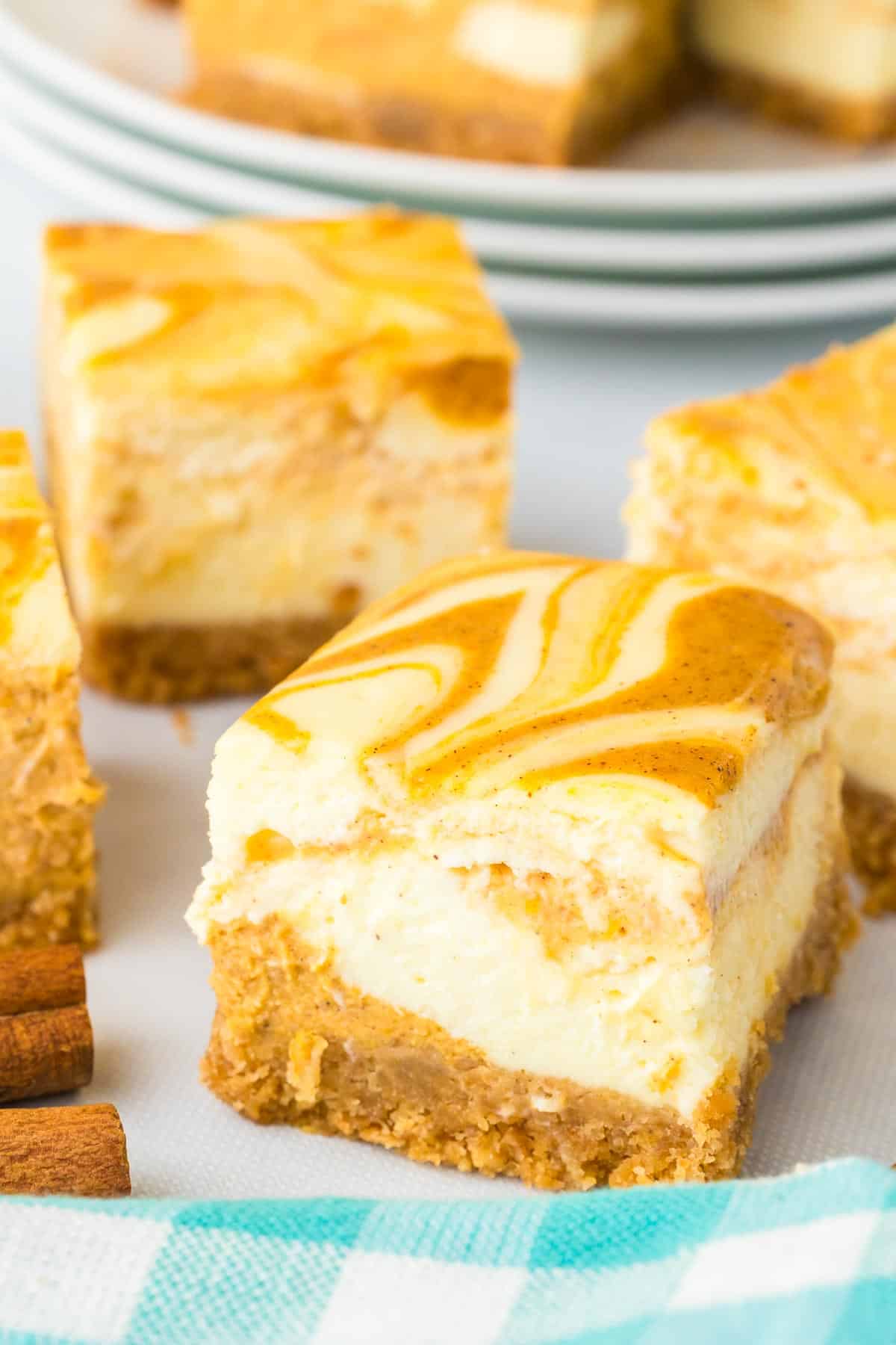 Pumpkin swirl cheesecake bars with a graham cracker crust on a white counter with more bars on a plate in the background.