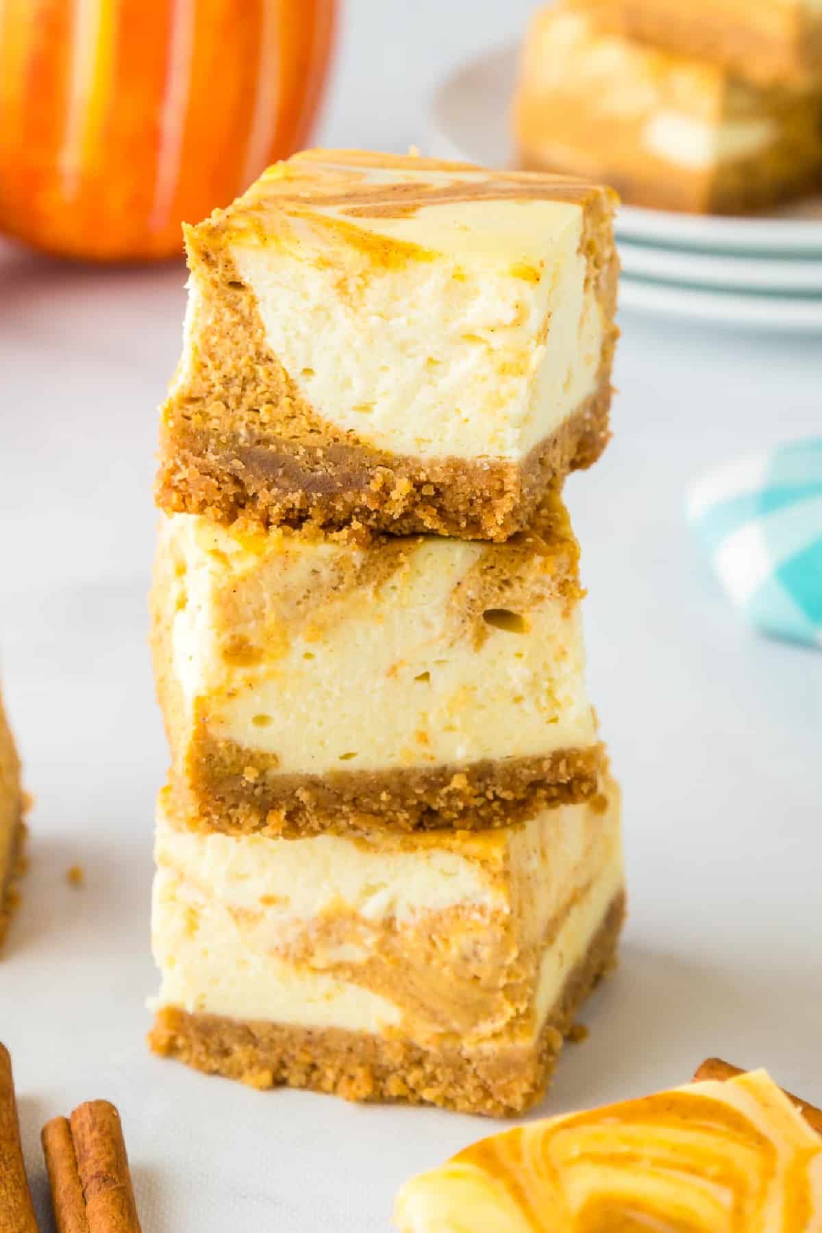 A stack of three pumpkin cheesecake bars with a graham cracker crust, in front of a whole pumpkin and plates.