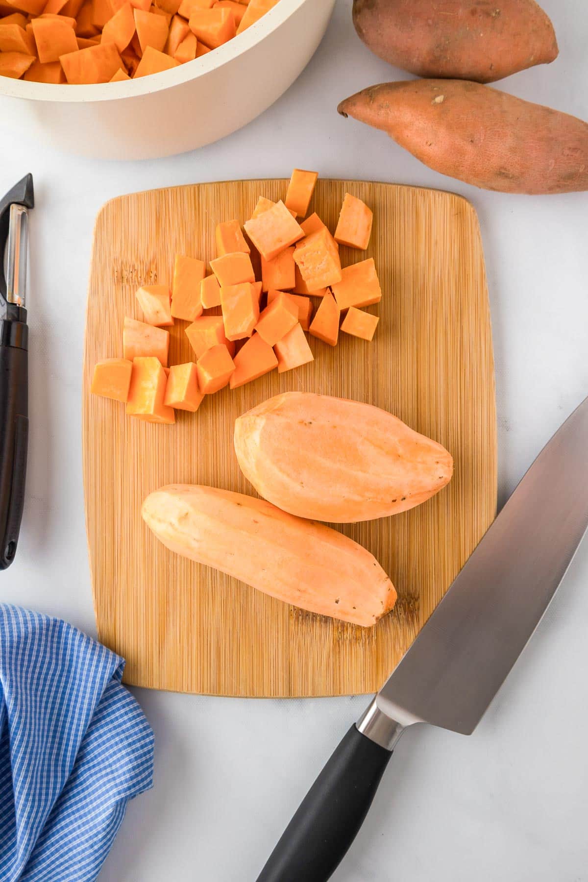 Diced and whole sweet potatoes on a wooden cutting board with a knife and a vegetable peeler for sweet potato casserole.