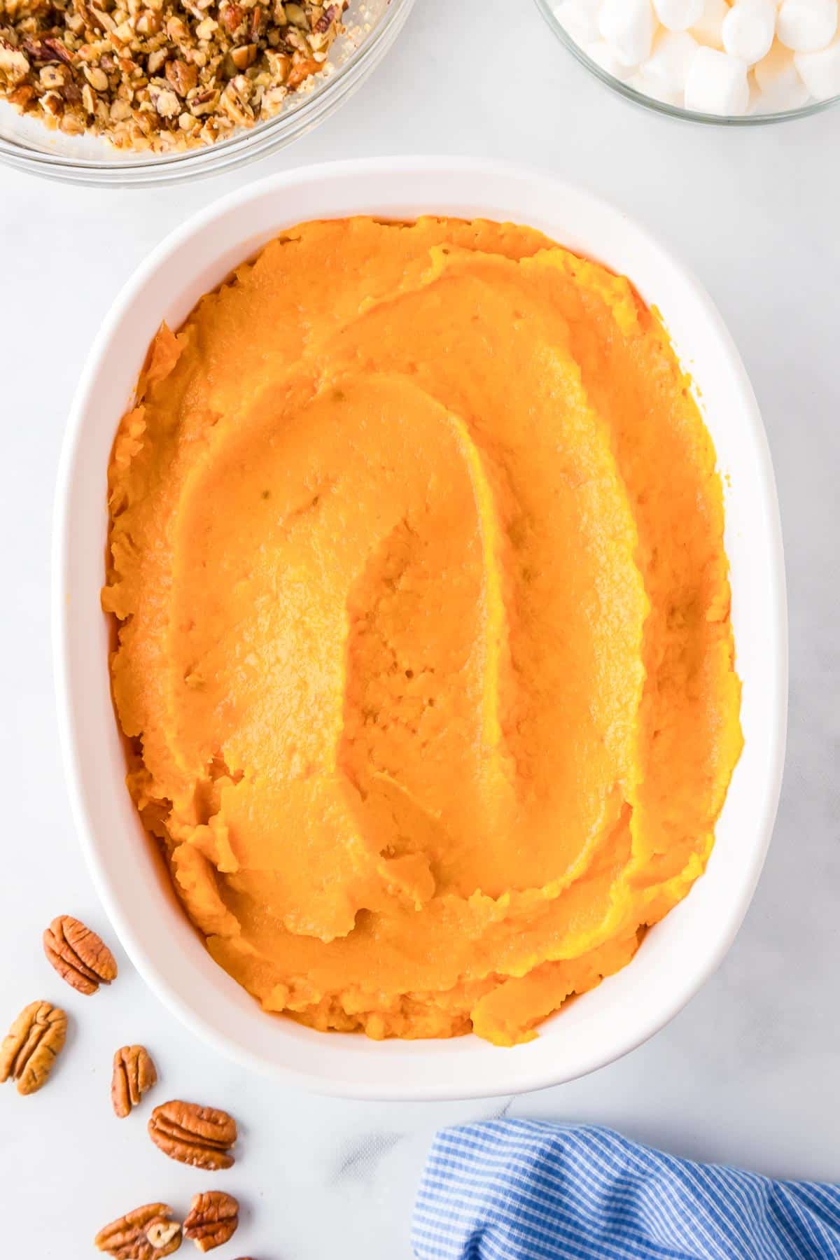 A baking dish full of mashed sweet potatoes with pecan topping and marshmallows in bowls nearby on the counter.