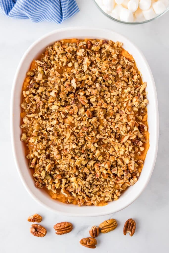 A baking dish full of sweet potato casserole topped with chopped pecans with marshmallows in a bowl on the counter nearby.