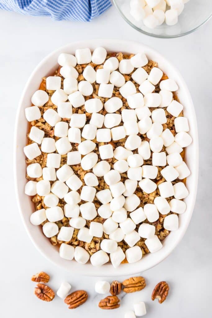 A baking dish filled with sweet potato casserole topped with marshmallows. Nearby are pecans and a bowl with additional marshmallows.