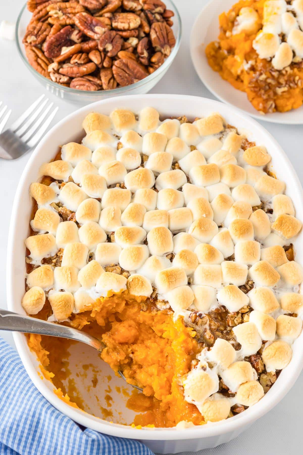 A casserole dish with sweet potato casserole topped with toasted marshmallows and pecans, with a serving spoon and a portion removed to a plate nearby and a bowl of pecans also nearby on the counter.