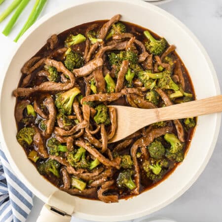 A square view of a skillet filled with stir-fried beef and broccoli in a dark sauce with a wooden spoon stirring.