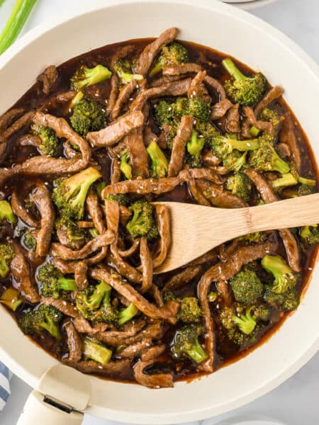 A square view of a skillet filled with stir-fried beef and broccoli in a dark sauce with a wooden spoon stirring.