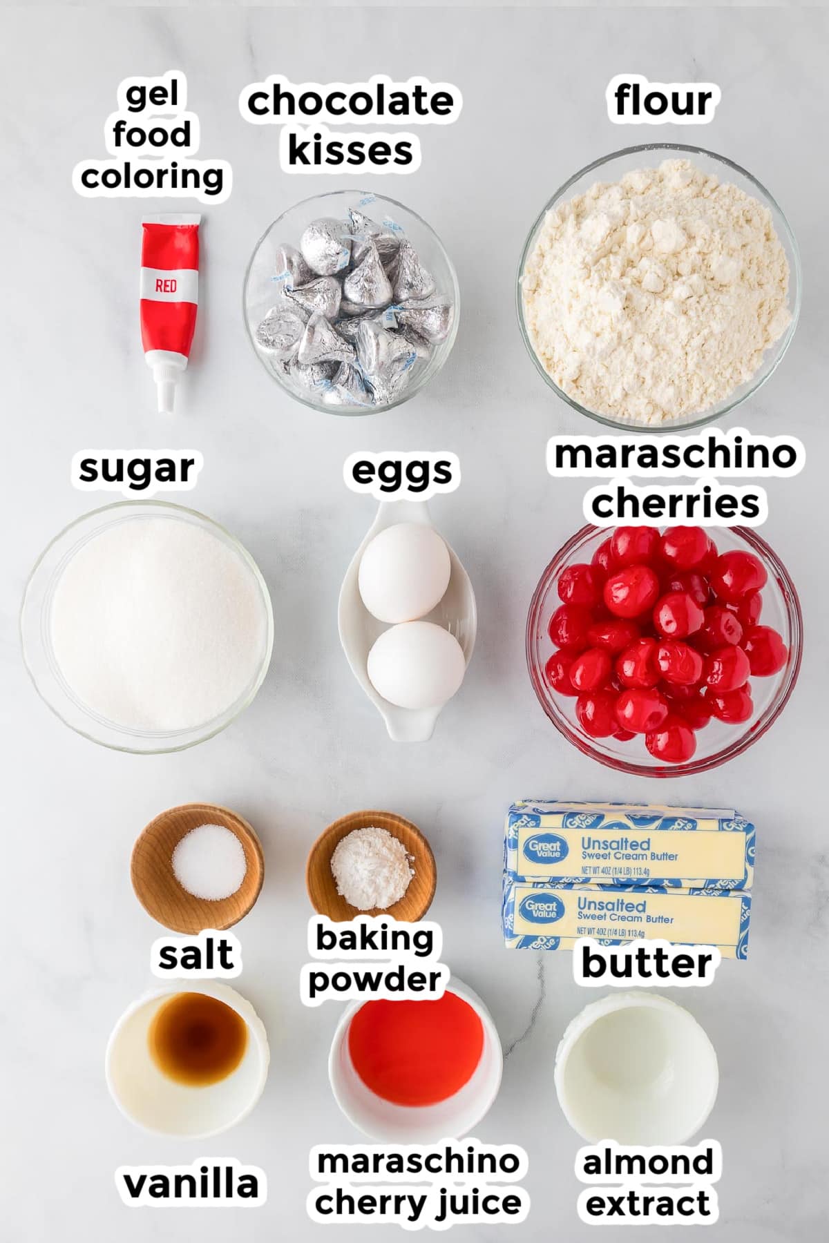 Labeled baking ingredients on a countertop, including flour, sugar, butter, eggs, chocolate kisses, maraschino cherries, maraschino cherry juice, vanilla, almond extract, baking powder, salt, and food coloring.