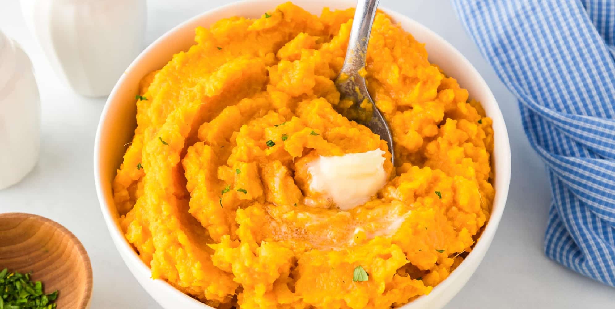 Wide view bowl of a bowl of maple mashed sweet potatoes with a pat of butter on top with a spoon scooping the sweet potatoes.