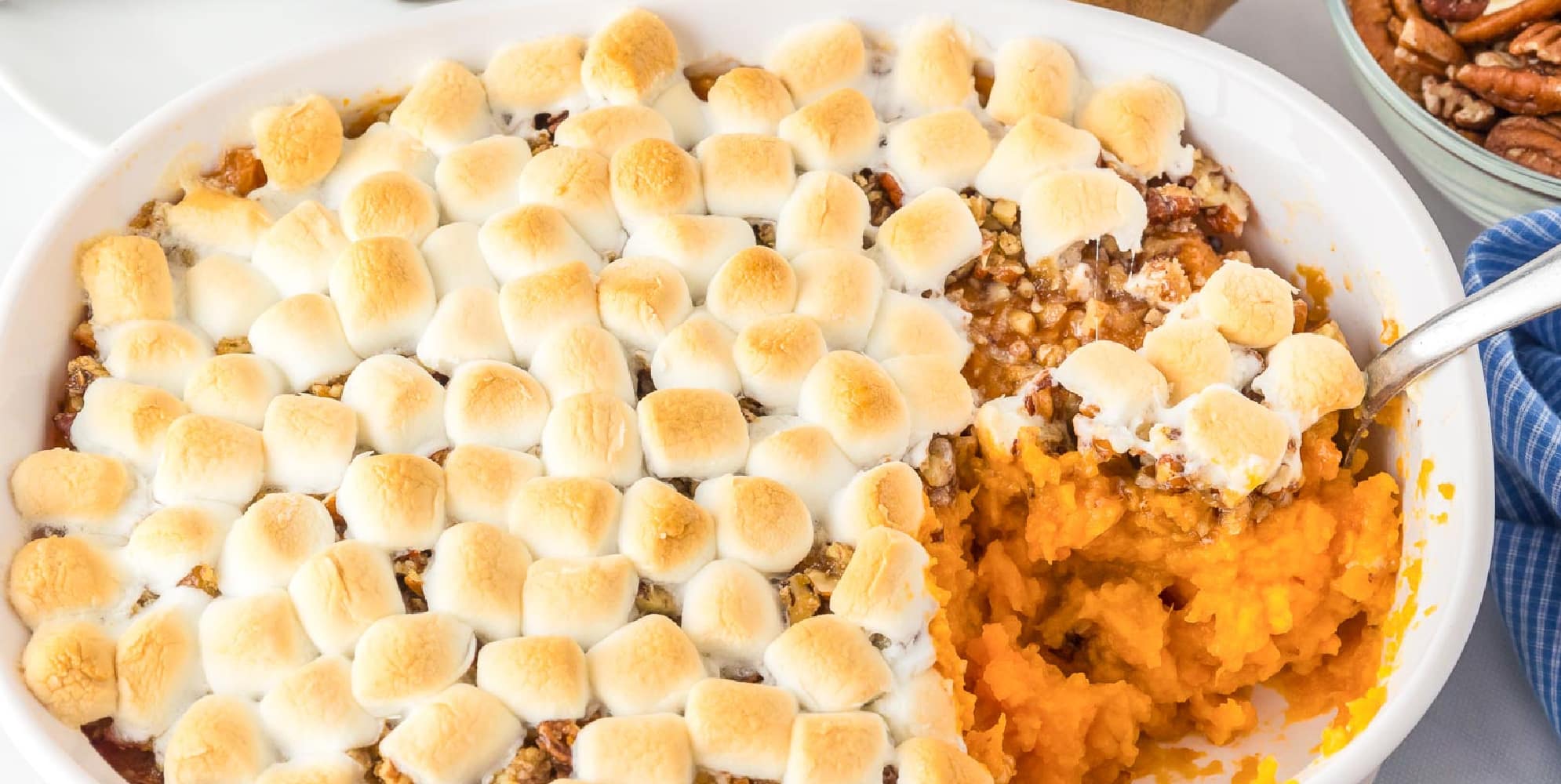 Wide view of a baking dish full of sweet potato casserole topped with toasted marshmallows and pecans, with a portion scooped out, revealing the mashed sweet potatoes underneath.