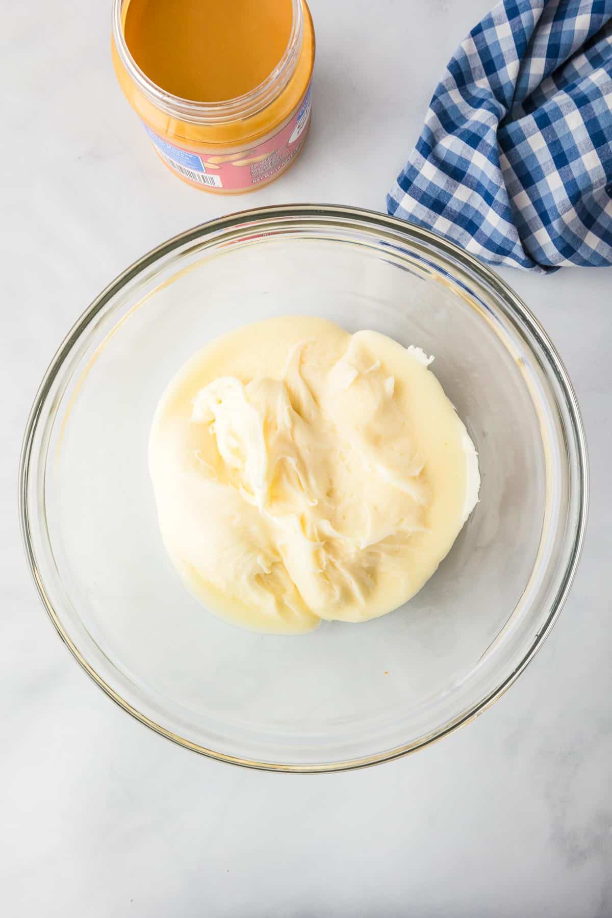 A glass bowl filled with melty cream cheese frosting with peanut butter nearby to make 2 ingredient peanut butter fudge.