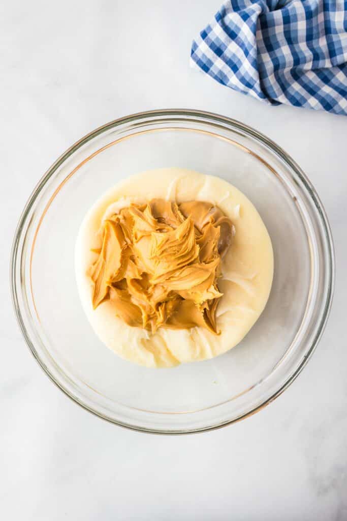 A glass bowl filled with a large amount of peanut butter and melty cream cheese frosting about to be stirred together for fudge.