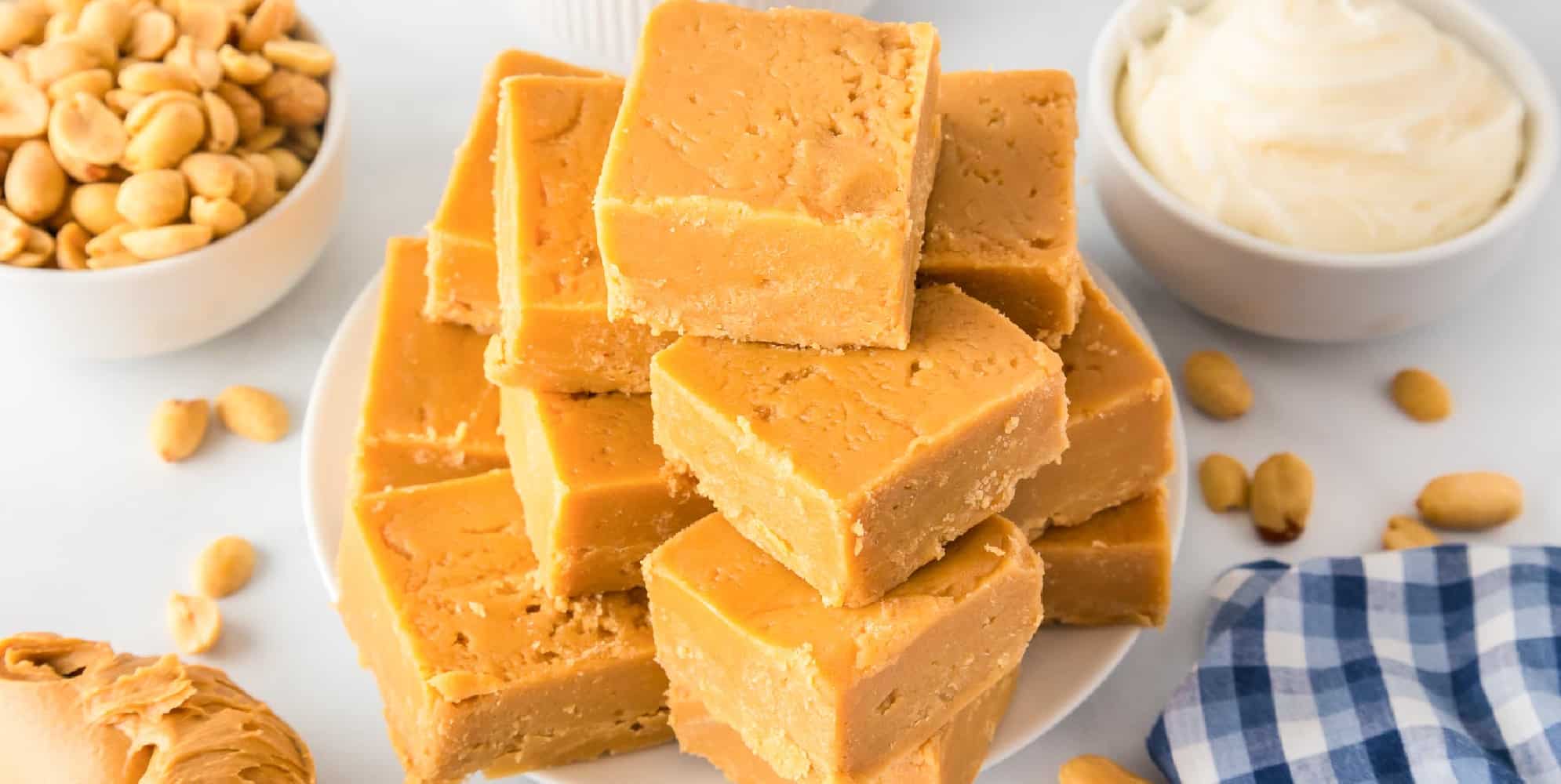 A wide view of a stack of 2 Ingredient peanut butter fudge piled high on a plate with more ingredients on the counter in bowls nearby.