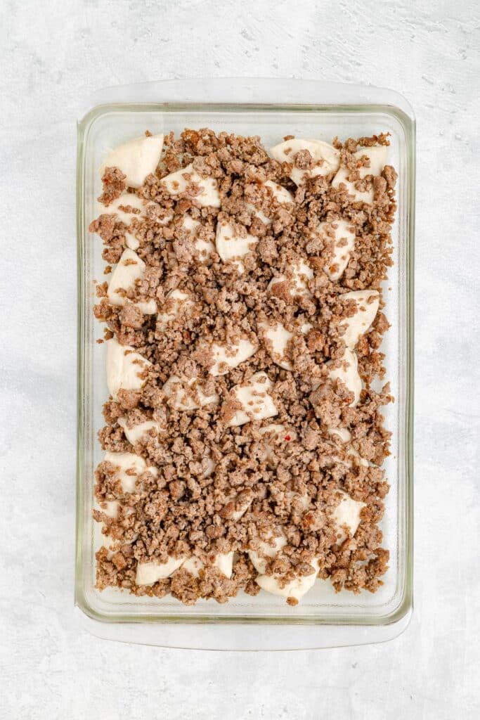 A glass casserole dish layered with biscuit dough and browned sausage for a breakfast biscuit casserole.