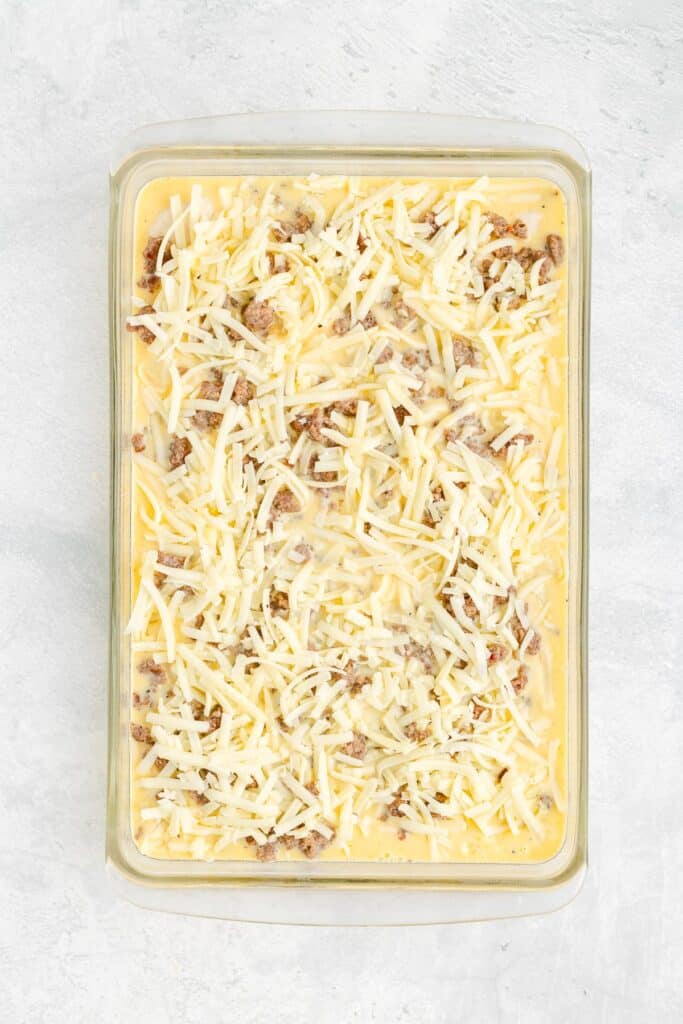 Glass casserole dish with breakfast biscuit casserole filled with a raw egg mixture, cooked sausage, biscuit dough pieces and shredded cheese.