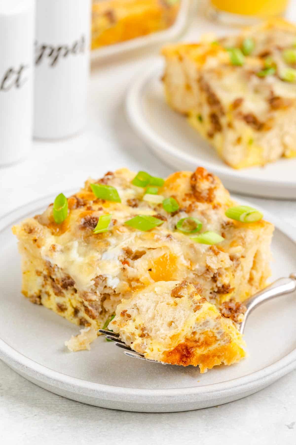 A slice of breakfast biscuit casserole on a plate with a fork removing a bite and another plate with a slice of casserole and more casserole in the pan in the background on the table.