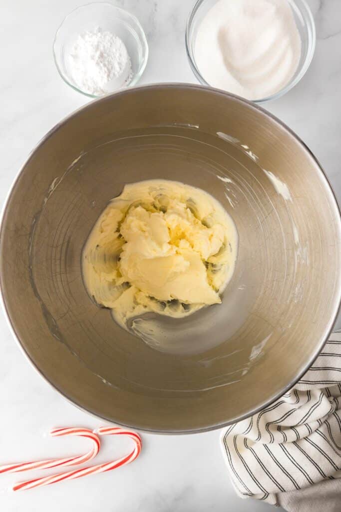 Softened butter in a large mixing bowl with sugar in bowls nearby on the counter for candy cane cookies.