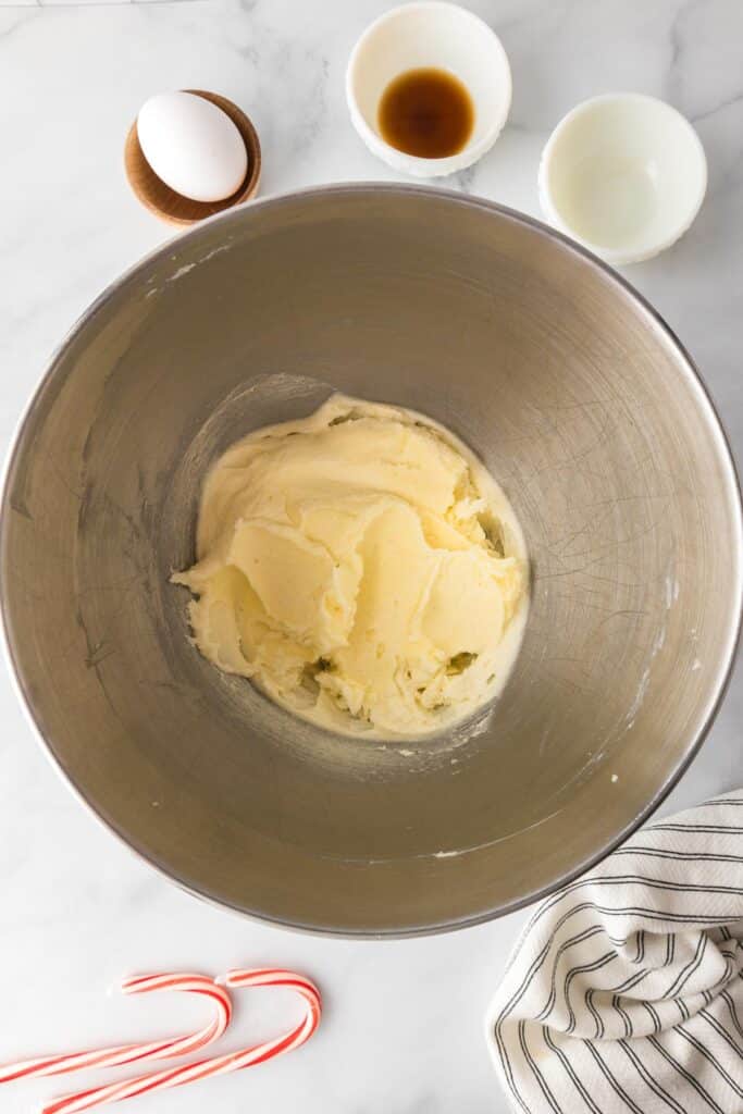 Mixing bowl with creamed butter and sugar on a countertop with vanilla extract, peppermint extract and an egg nearby on the counter for candy cane cookies.