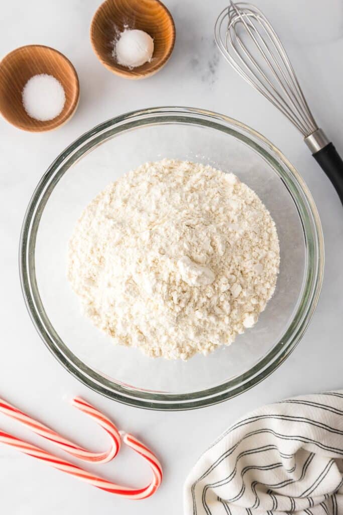 A large mixing bowl with flour on a countertop with bowls of salt and baking powder and a whisk nearby on the counter.