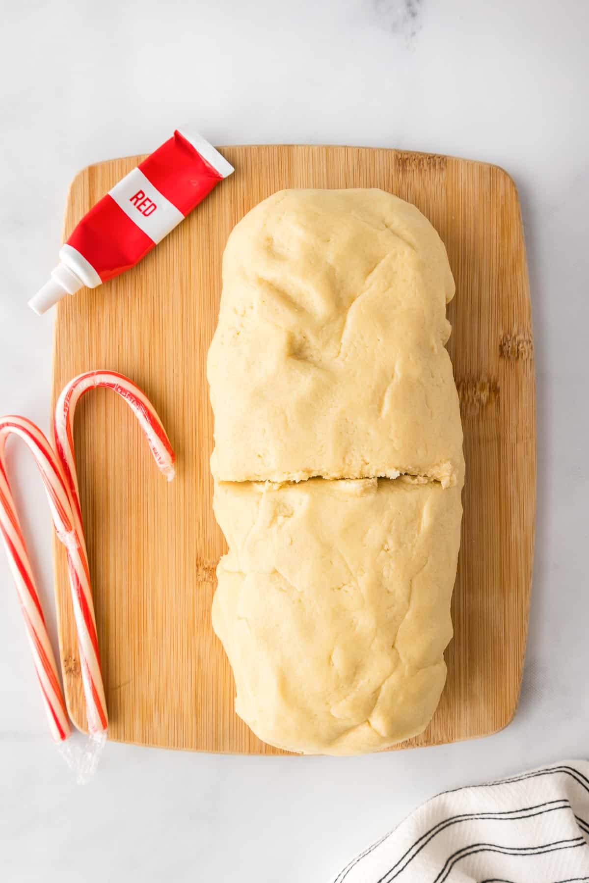 A block of cookie dough on a wooden cutting board a red food coloring tube and two candy canes nearby.