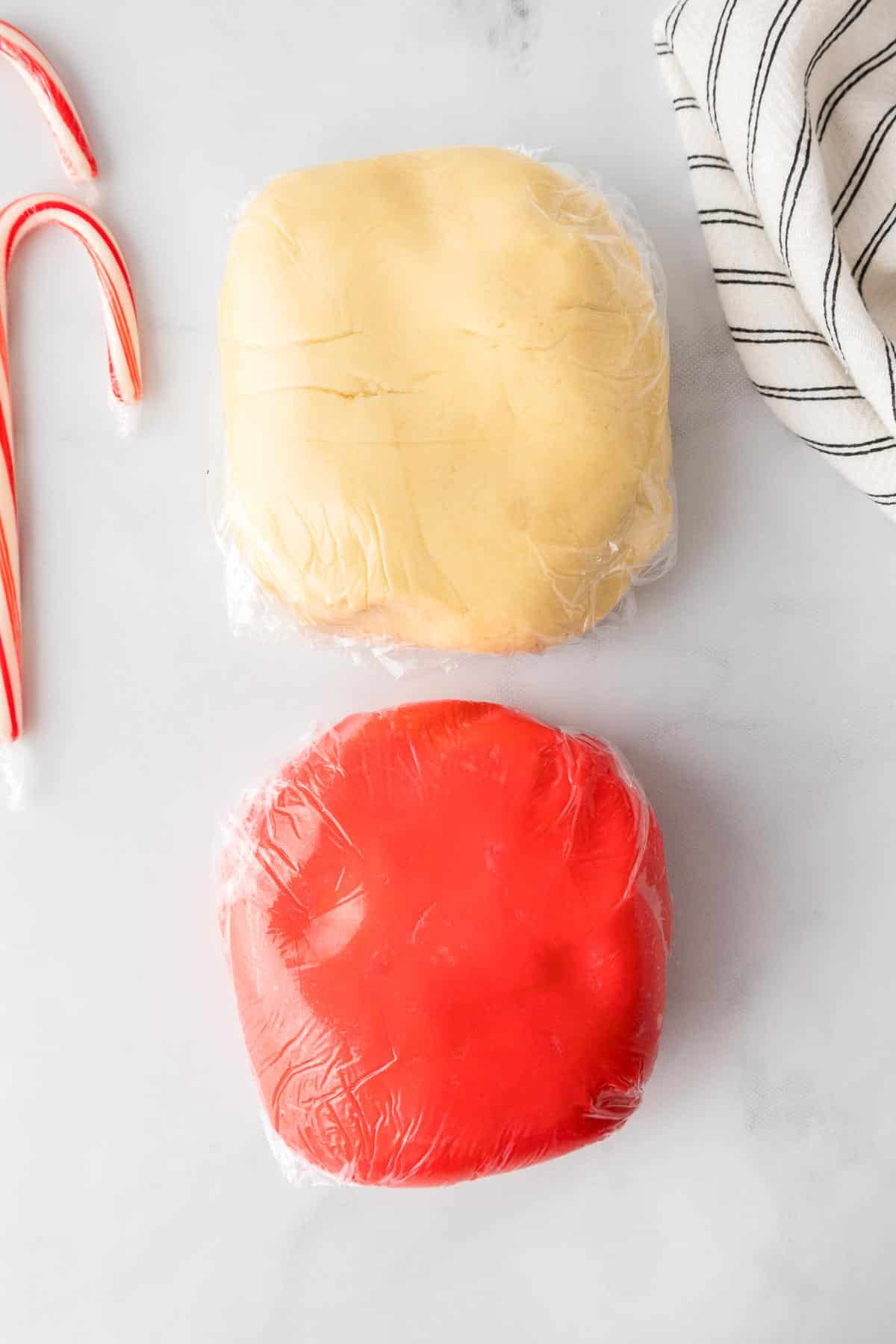 Two blocks of cookie dough, one red and one un colored, wrapped in plastic wrap.