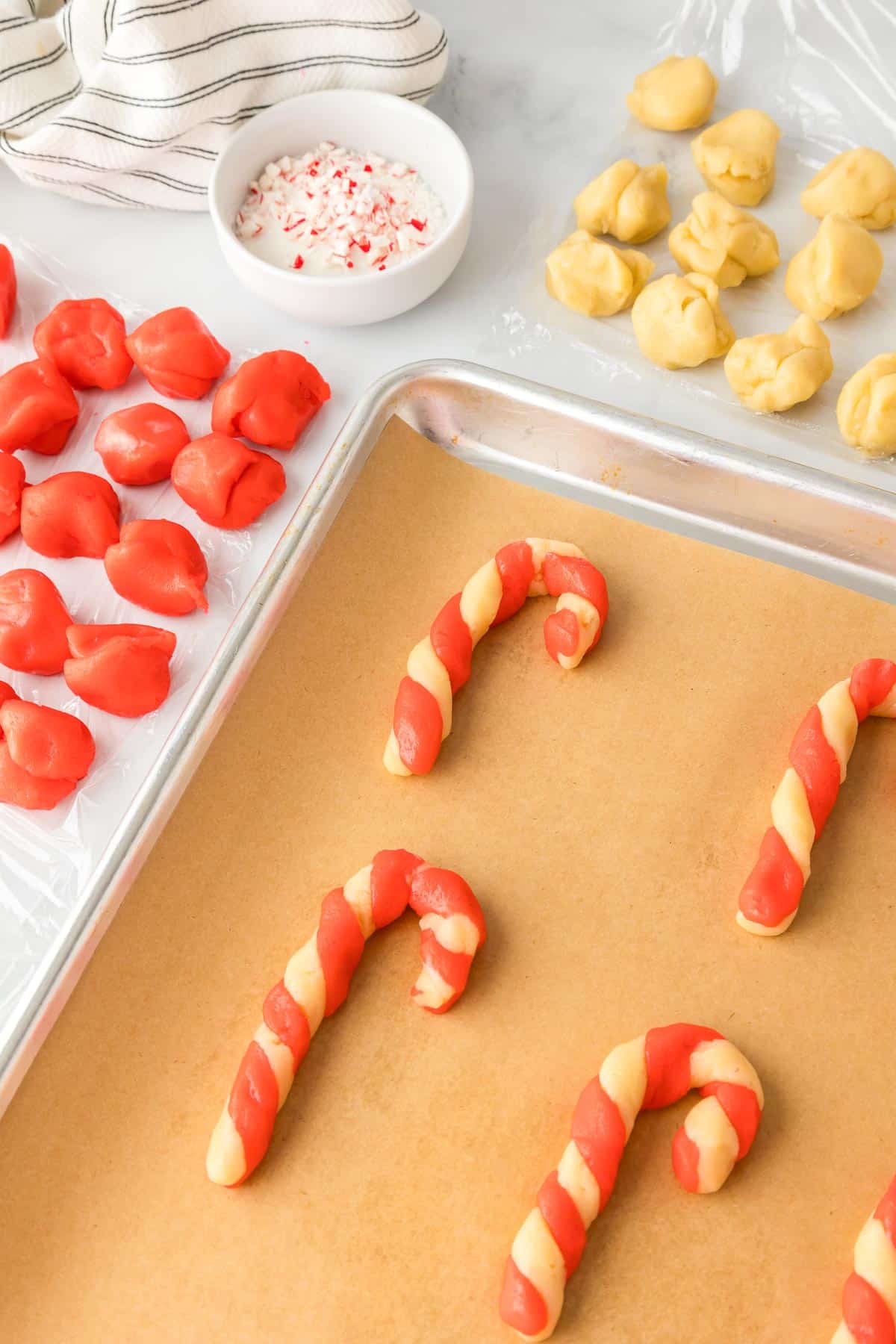 Unbaked candy cane cookies on a baking pan with dough balls and crushed peppermint in a bowl the background.