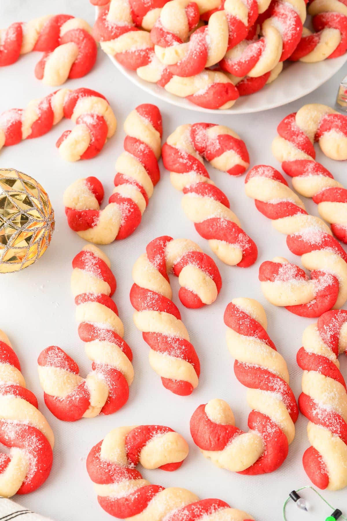 Many red and white candy cane shaped cookies on a counter with a plate full of more candy cane cookies and a few holiday decorations nearby.