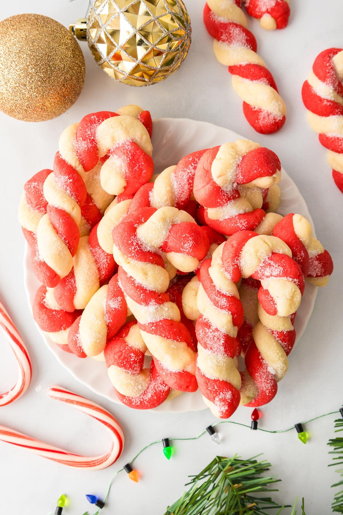 A plate full of red and white shaped candy cane cookies, surrounded by Christmas decorations nearby on a counter.