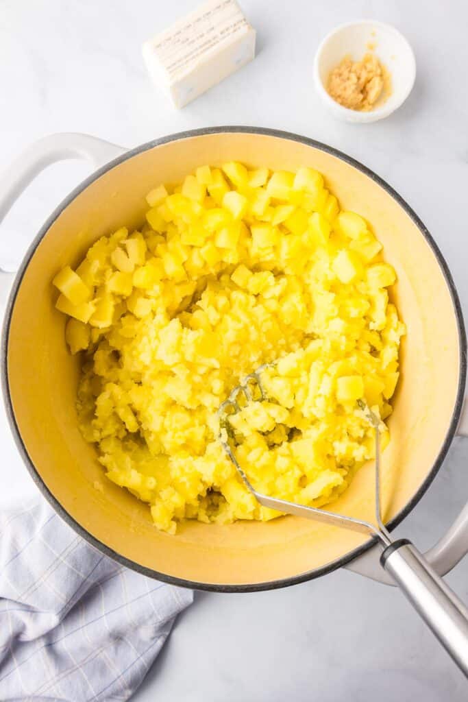 A pot of partially mashed yellow potatoes being mashed with a potato masher with butter and garlic nearby on the counter.