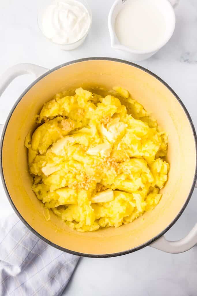 A pot of yellow Yukon gold mashed potatoes with butter and garlic on top, with milk and sour cream nearby on the counter.