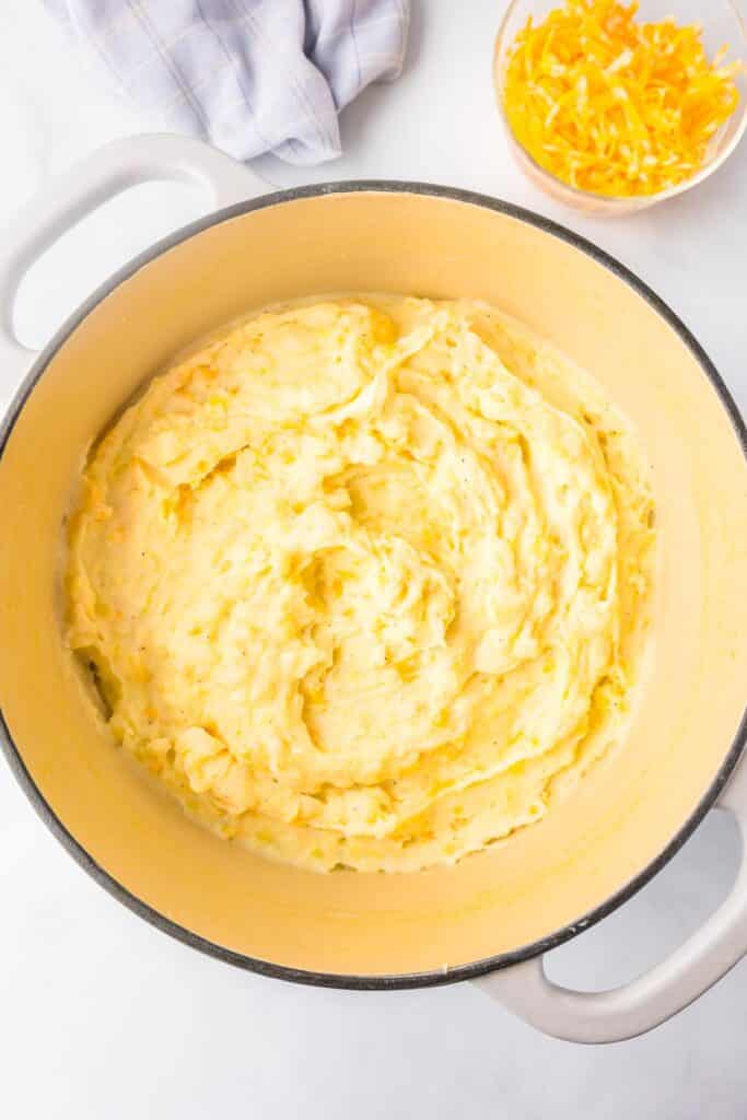 A pot filled with creamy yellow yukon gold mashed potatoes on a counter top with cheese in a bowl nearby.