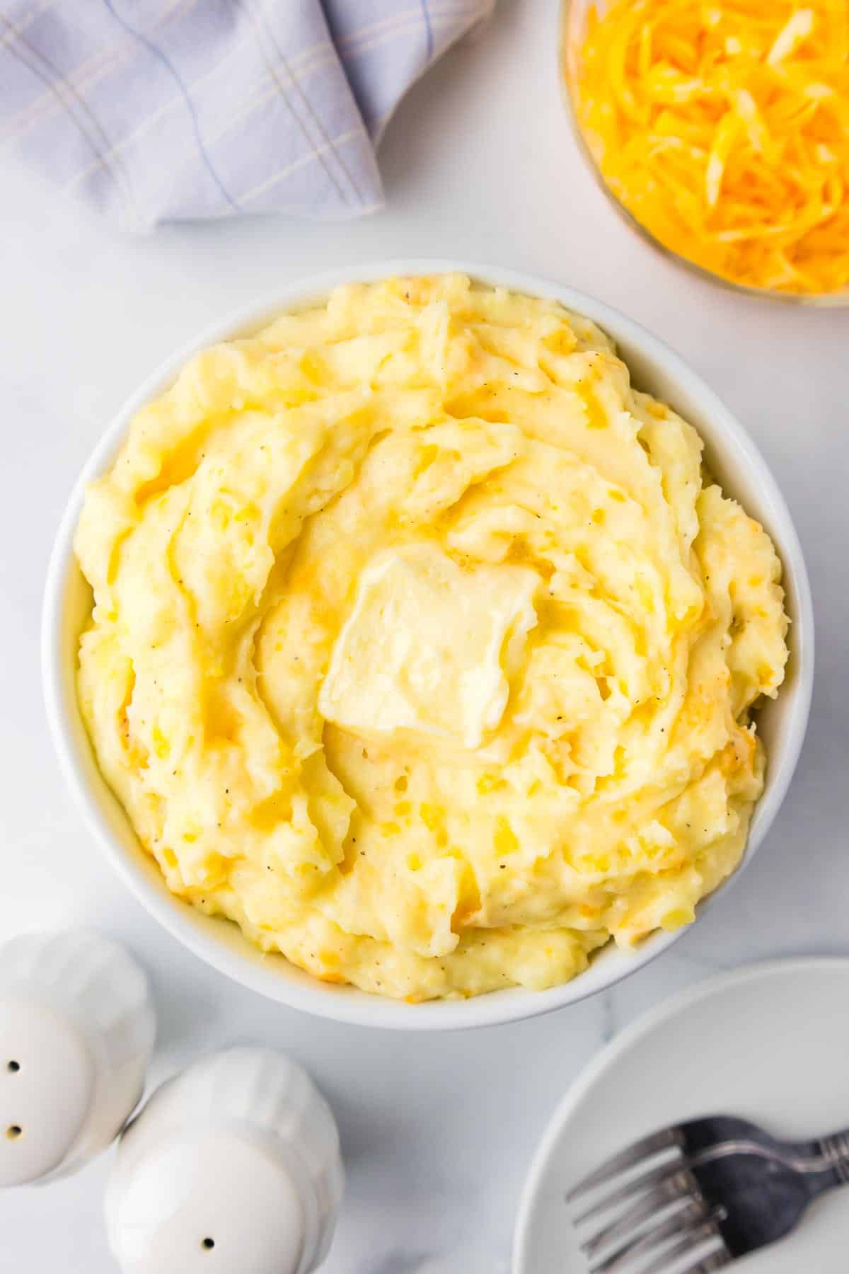 A bowl of mashed Yukon Gold potatoes topped with a melty pat of butter with a bowl of shredded cheese, plates stacked with forks, and salt and pepper shakers nearby.