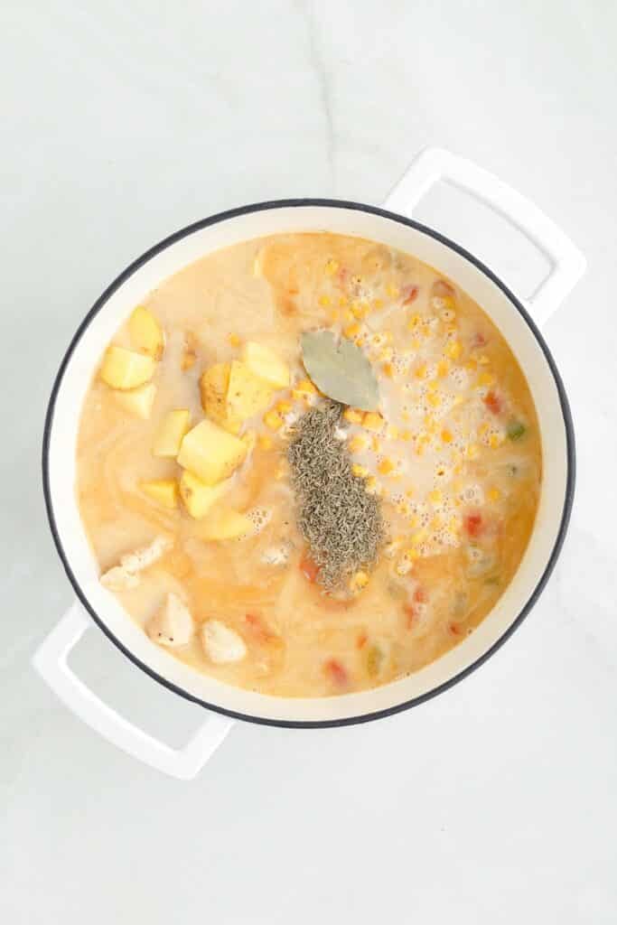 A pot filled with a creamy chicken corn chowder soup containing diced potatoes, vegetables, cooked chicken, a bay leaf, and a sprinkle of herbs.