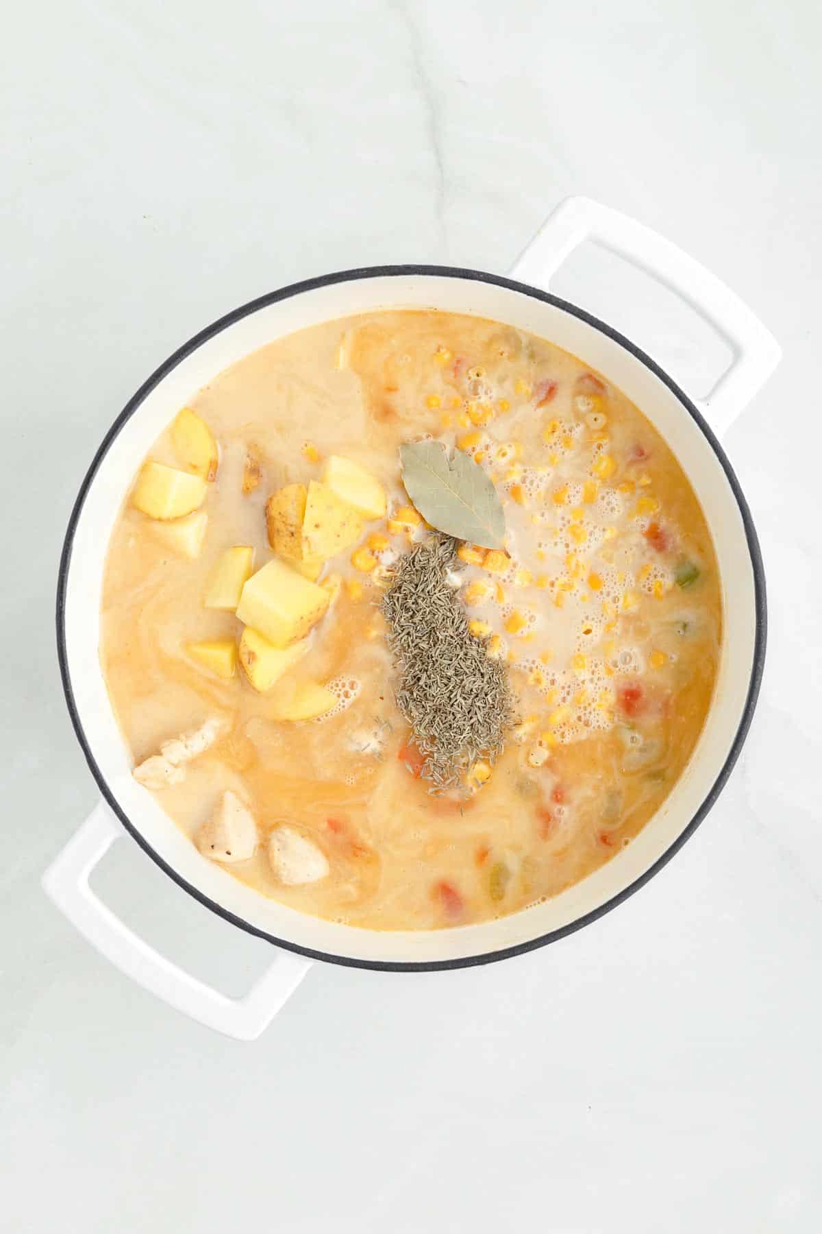 A pot filled with a creamy chicken corn chowder soup containing diced potatoes, vegetables, cooked chicken, a bay leaf, and a sprinkle of herbs.