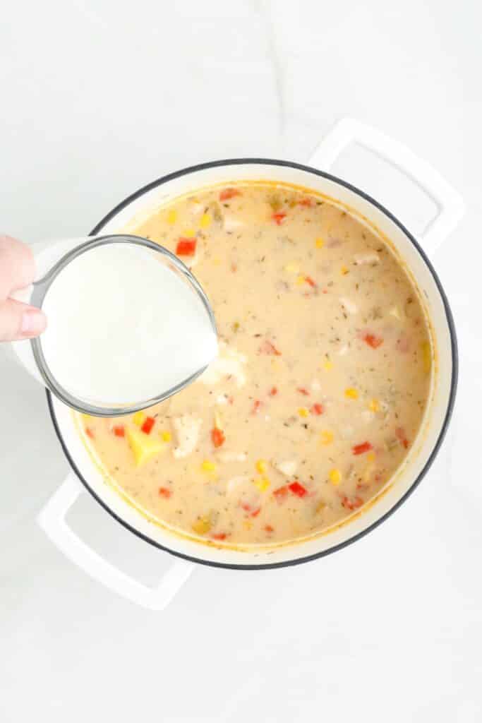 A hand pours a measuring cup full of heavy cream into a pot of chicken corn chowder.