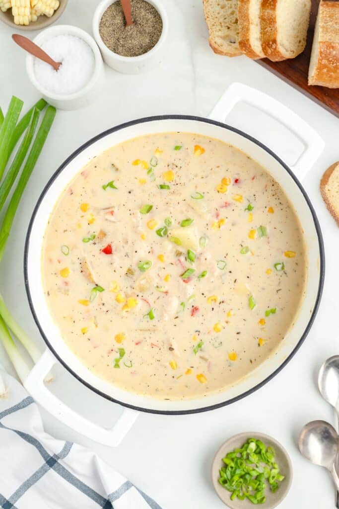 A pot of creamy chicken corn chowder oun a counter garnished with chopped green onions surrounded by bread, a napkin, spoons being served on a table.